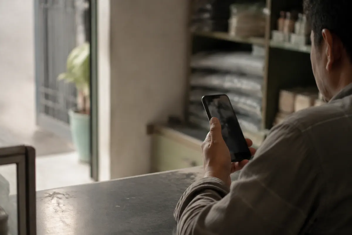 Alguien revisa su celular apoyado en el mostrador de un negocio pequeño, con los estantes desenfocados al fondo